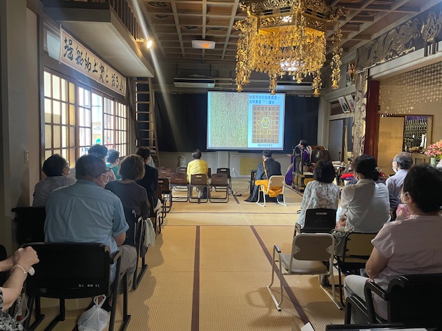 読経前の「福性寺の歴史」講演 スクリーンの左か光明真言塔の光明真言と胎蔵界大日如来真言 　右は福性寺本堂の天井の光明真言